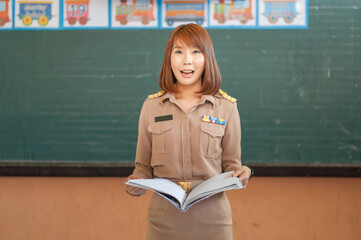 female thai teacher in government uniform standing in front of classroom reading book to class