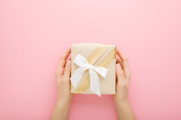 Young adult woman hands holding beige paper gift box with white ribbon on light pink table background. Pastel color. Closeup. Point of view shot. Congratulation concept. Top down view.