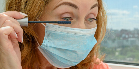 An adult red-haired woman in a medical mask applies mascara to her eyelashes. Selective focus. Close up.