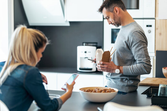 Serious And Unhappy Young Couple Eating While Using Smartphone In The Kitchen At Home.