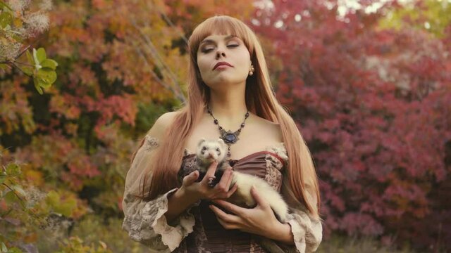 portrait beautiful redhead woman forest nymph with pet ferret on shoulder. fashion model in medieval dress brown beige clothes. background Autumn nature trees orange red leaves. Girl fantasy princess