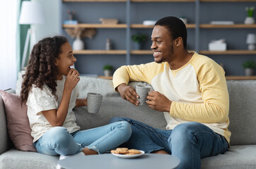 Cute black father and daughter drinking tea with cookies