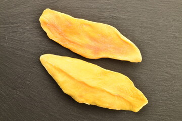 Two light yellow dry mango slices on a slate board, close-up, top view.