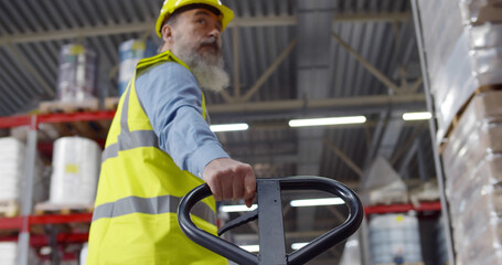 Senior worker in warehouse using hand pallet stacker to transport goods.