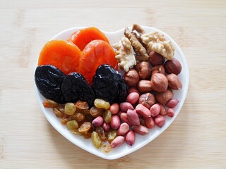 Healthy breakfast. Heart shaped white plate with dried fruits on wooden background. Dried apricots, prunes and nuts. Healthy food and snack. Flat lay. Place for text