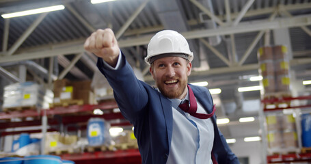 Portrait of superhero businessman in hardhat and formal suit in warehouse