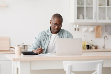 Budget planning concept. Pensive african american man calculating taxes and using laptop computer, sitting in kitchen