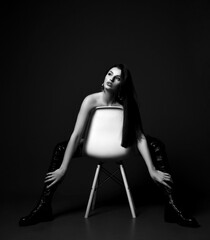 Black and white. Gorgeous sexy woman vamp naked and in high leather boots sits backwards on white chair over dark background. Fashion, vogue, sexy stylish look for woman concept