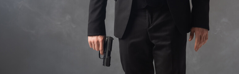 partial view of businessman in black suit holding weapon on grey background with smoke, banner