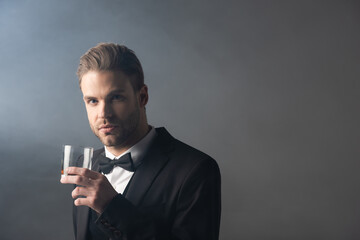 successful businessman looking at camera while holding glass of whiskey on grey background with smoke