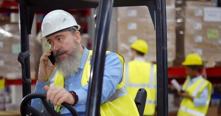 Senior forklift driver sitting in vehicle in warehouse talking on smartphone