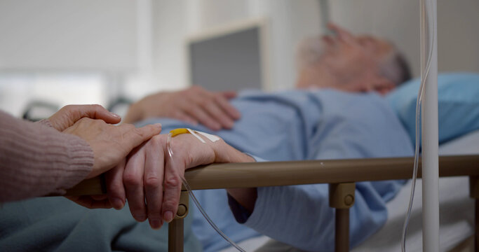 Sick Senior Man In Hospital Bed And His Wife Sitting Next To Him