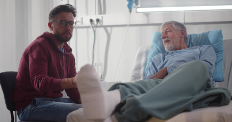 Adult man visiting aged father with broken leg in hospital ward