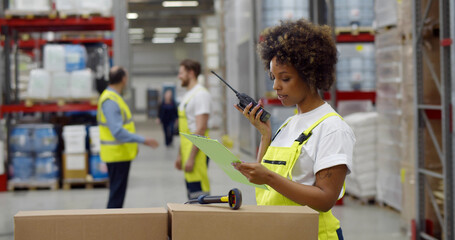 African female warehouse manager holding clipboard and using walkie-talkie while checking inventory.