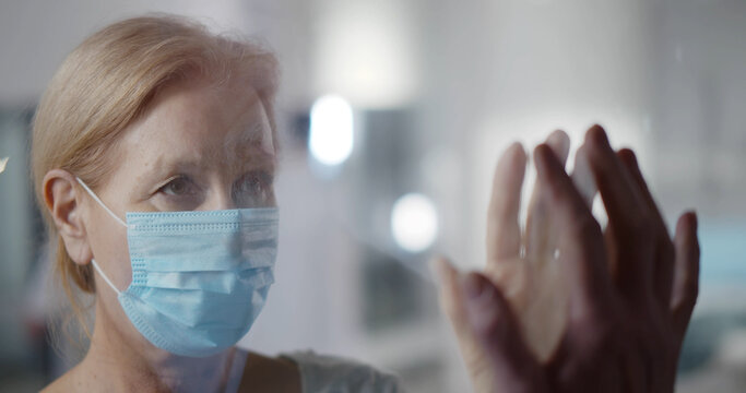 Sick Aged Woman In Safety Mask Talking To Family Visitor Through Protective Glass Wall In Hospital.