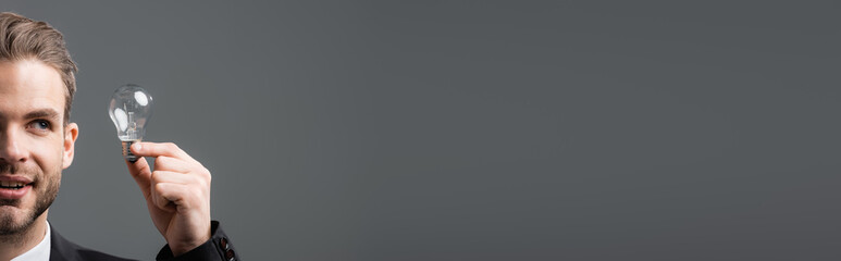 partial view of smiling businessman looking at light bulb isolated on grey, banner