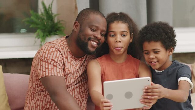 Medium closeup of cute afro siblings age 8 and 10 taking selfie with their funny dad on digital tablet, sitting together on comfy couch in living room