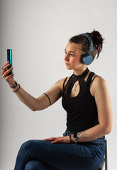 Alternative girl with tattoo using her cell phone and wireless headphones wearing a sleeveless top. Person in a video meeting.