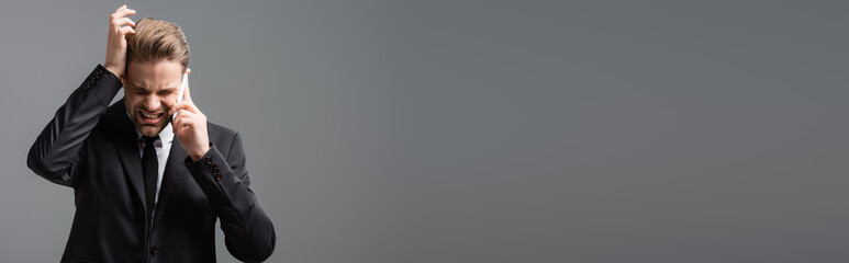 depressed businessman touching head while calling on smartphone isolated on grey, banner