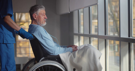 Nurse putting wheelchair with senior man with nasal oxygen tube near window