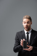 surprised businessman holding mobile phone with blank screen isolated on grey