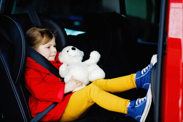Toddler girl sitting in car seat, holding plush soft doll toy and looking out of the window on traffic. Little kid traveling by car. Child safety on the road. Family trip and vacations