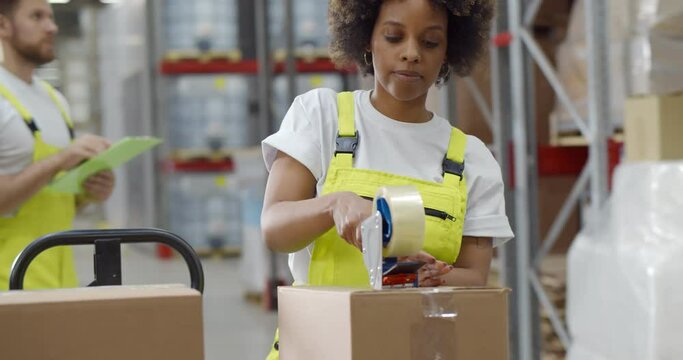 African female worker packing cardboard box with tape gun dispenser in warehouse