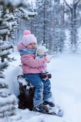 Fototapeta na wymiar Toddler little girl sits on a stump in snowsuit on a winter day. hugs a knitted toy