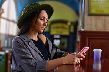 Stylish fashionable elegant cute attractive hipster woman traveler wearing felt hat using a phone during resting at cafe shop