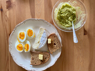 Breakfast Plate with Brie Cheese, Mashed Avocado, Boiled Eggs and Butter Bread on Wooden Table.
