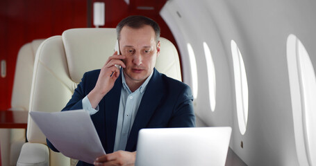 Mature businessman talking on phone and holding document sitting in private business plane