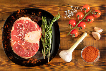 Raw meat with a bone on a black plate. Cherry tomatoes, garlic, spices. Wooden table. Top view.