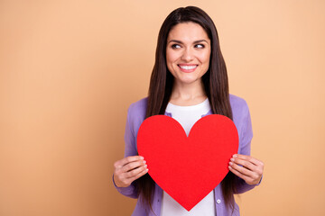 Photo of cheerful woman happy smile dream look empty space hold paper heart love romance isolated over beige color background