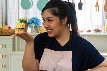 Unhealthy obesity with sweet cake or weight loss concept. Happiness young woman wear apron holding candy cupcake in hands while sitting in the kitchen at home.