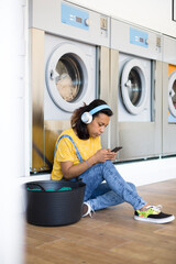 Young hispanic woman sitting on the floor listening to music and using her smartphone. She is waiting for her clothes to be washed at a self-service laundry.