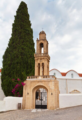 Fototapeta na wymiar Greece. Rhodes Island. Church of St. John the Evangelist in the village of Vati with a beautiful old stone bell tower and a cypress tree at the entrance on a summer day. Summer travel and excursions