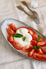 Traditional Italian salad with Burrata cheese, tomatoes and fresh basil. Mozzarella burrata salad with basil leaves and tomatoes on plate on beige linen tablecloth
