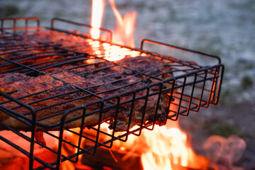 Cooking chicken meat over charcoal. Chicken meat fried with a crust on a wire rack during grill cooking. Backlight from a burning fire and smoke in the late evening.