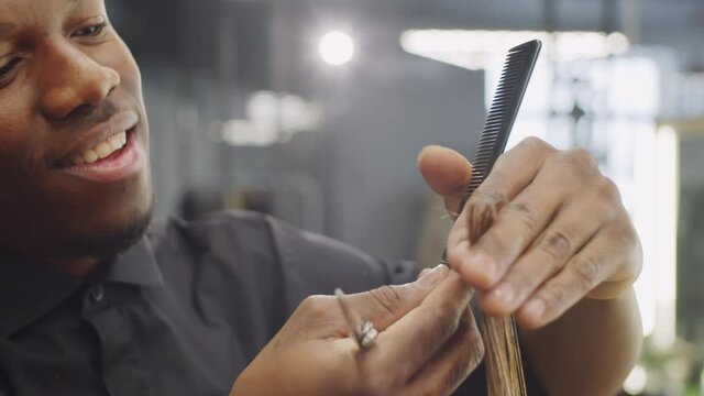 Close up shot of young handsome black barber smiling and talking while cutting hair of client with shears and comb in barbershop