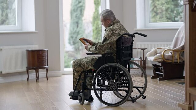 Wide shot of upset frustrated disabled man in wheelchair looking at picture sitting at home indoors. Depressed Caucasian retired veteran alone. Social issues and loneliness concept
