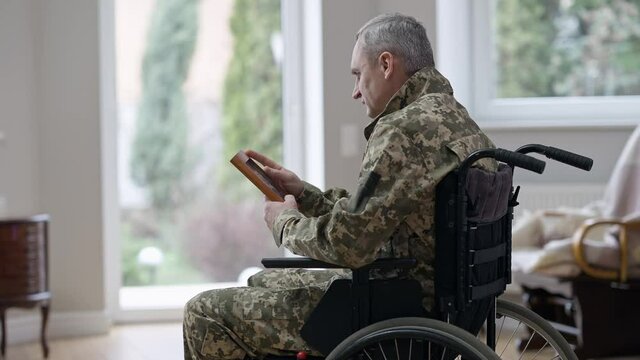 Side view of frustrated middle aged man in wheelchair looking at picture and crying. Depressed disabled Caucasian middle aged veteran recalling memories at home indoors. Handicap and frustration