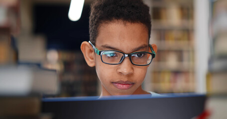Close up of preteen african boy student smiling typing on laptop working at school library.