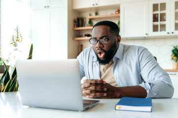 Shocked amazed African American man with glasses, freelancer, ceo or stock agent sits at his desk, looking at laptop in surprise, reading unexpected news or got big account profit