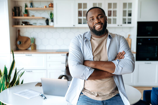 Friendly handsome confident stylish bearded African American male freelancer, standing near the workplace at home, crossing his arms, looking at the camera smiling friendly. Work from home concept