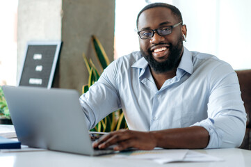 A portrait of an attractive good looking african american man, real estate agent, lawyer or ceo, manager, wearing formal stylish clothes and glasses, sits at workplace, looks at camera, friendly smile