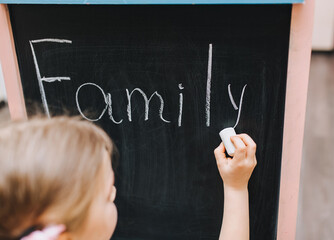 Little girl writes on a wooden board, easel with white chalk the word family. Home school assignment.
