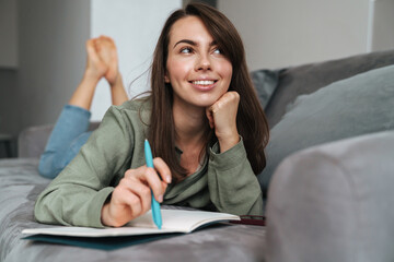 Young smiling woman writing down notes while lying on sofa at home