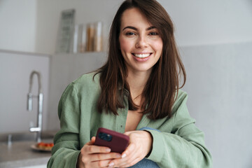 Young smiling woman using mobile phone while sitting at home kitchen
