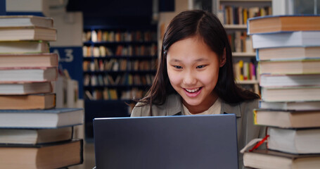 Asian schoolgirl studying on laptop and smiling sitting in school library