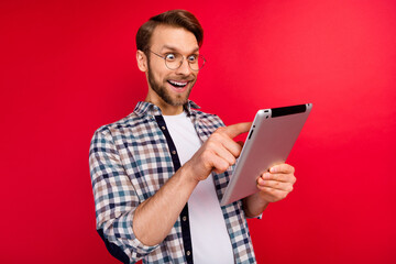 Photo of excited funny young guy wear checkered shirt glasses typing modern device big eyes isolated red color background